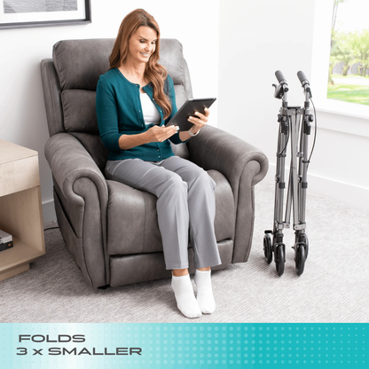 A woman reads a tablet in a gray recliner, with the Stander Wonder Indoor Rollator with Tray Table folded nearby. Text at the bottom says, FOLDS 3x SMALLER. Bright room and large window in the background.
