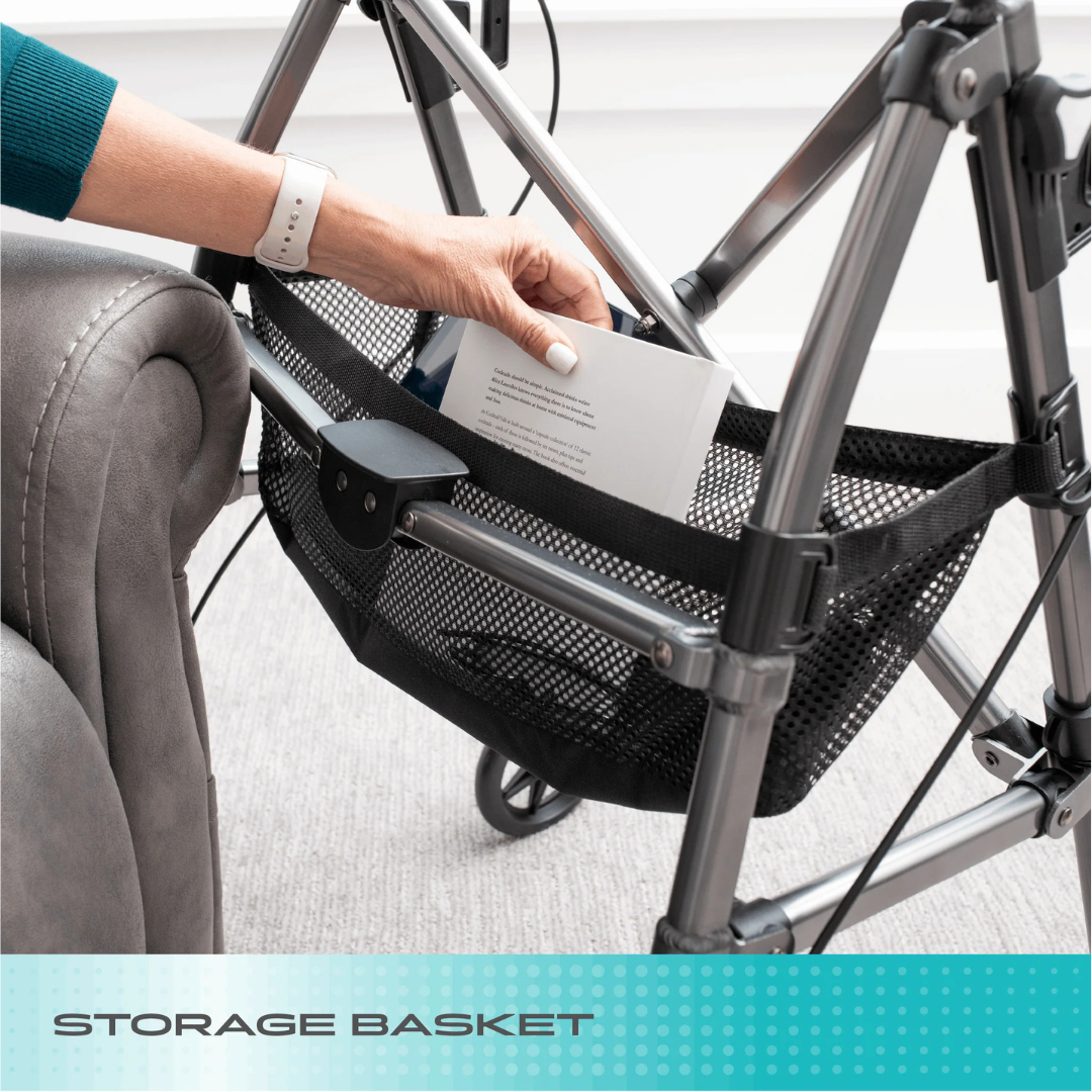 A hand places a book into the black mesh storage basket of the Stander Wonder Indoor Rollator with Tray Table, positioned beside a gray armchair. The text “Storage Basket” appears at the bottom.