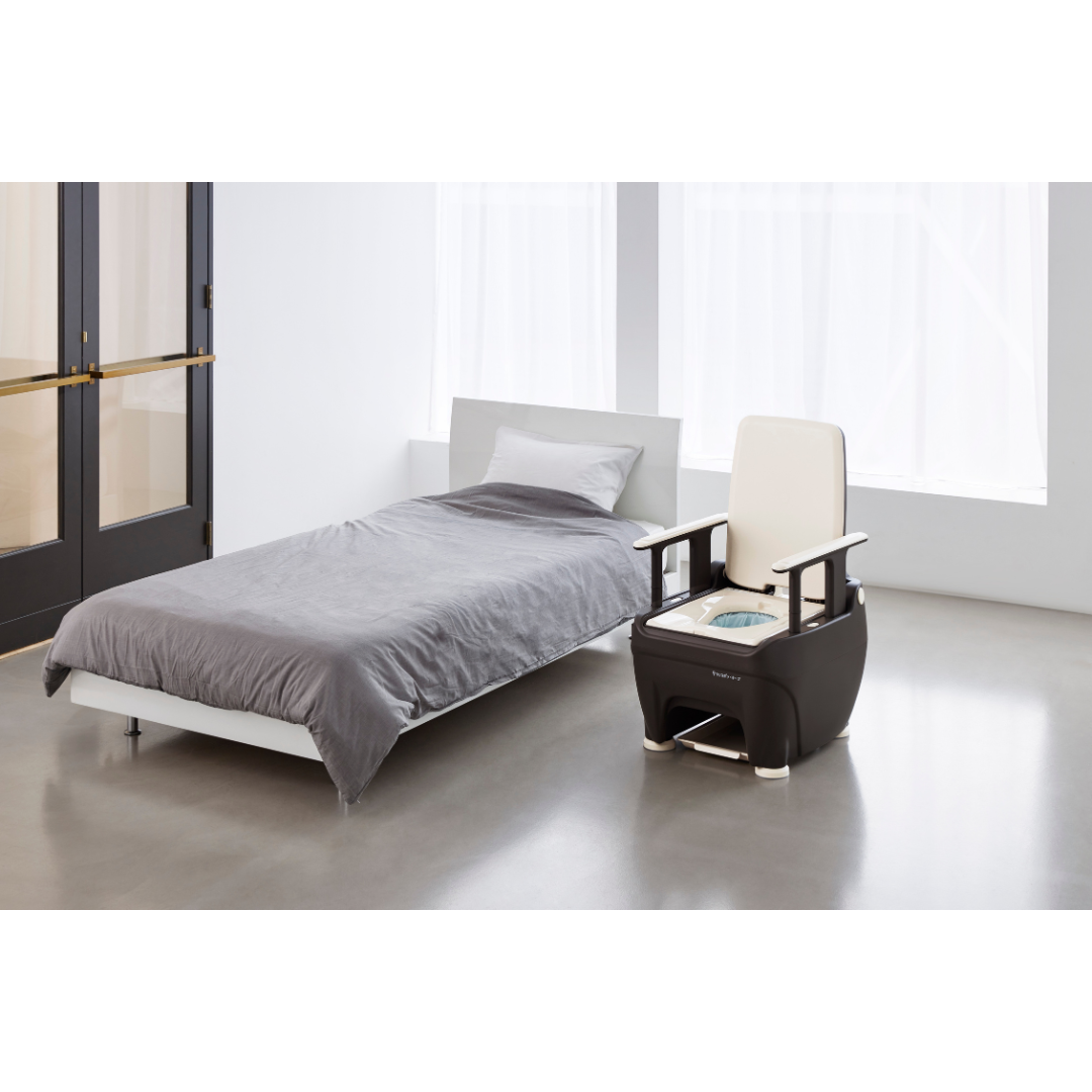 A minimalist bedroom with a low bed in gray bedding and the Wrappon® ORB Portable Commode Toilet with auto waste sealing, featuring built-in controls, in a bright room with large windows and a glass door.