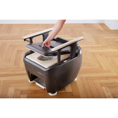A person lifts the lid of a portable dark gray Wrappon® ORB - Portable Commode Toilet with Auto Waste Sealing, which has wooden armrests and is placed on a wooden floor in a bright room, revealing its inner compartment.