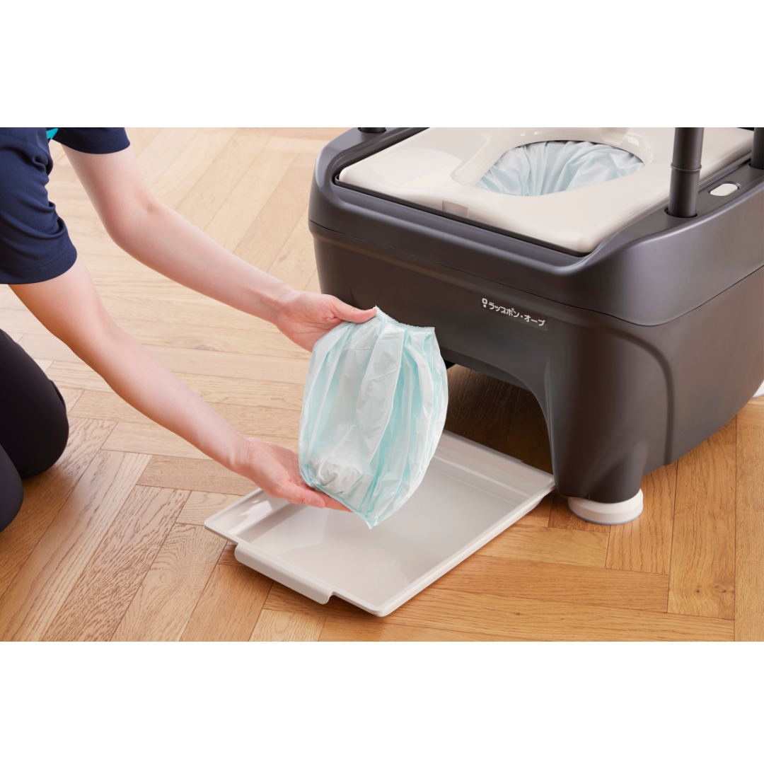 A person kneels on a wooden floor, removing a sealed light blue waste bag from the Wrappon® ORB - Portable Commode Toilet with Auto Waste Sealing, which features a black and white pull-out tray.
