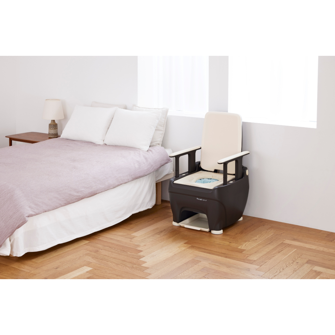 A bedroom with a neatly made bed, a wooden nightstand with a lamp, and a modern Wrappon® ORB - Portable Commode Toilet with Auto Waste Sealing placed on the wooden floor near a window.