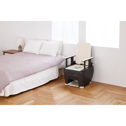 A bedroom with a neatly made bed, a wooden nightstand with a lamp, and a modern Wrappon® ORB - Portable Commode Toilet with Auto Waste Sealing placed on the wooden floor near a window.