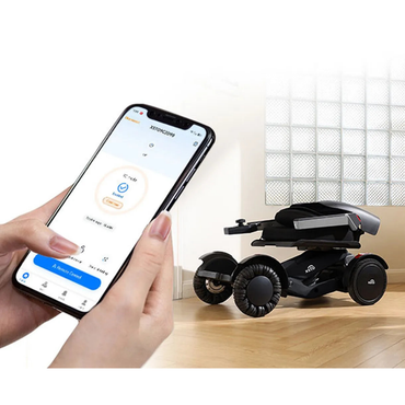 A person uses a smartphone app to control the XSTO Mobility M4 Self-Balancing Powerchair—a compact, black, four-wheeled electric wheelchair—on a light wooden floor near a window, highlighting its lightweight, foldable design for home or travel.