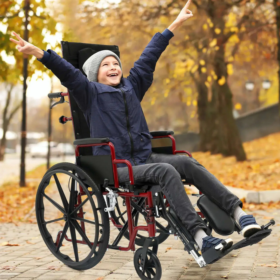 A smiling boy raises his arms joyfully outdoors in autumn, seated in the Circle Specialty ZIGGO Pro Reclining Pediatric Wheelchair. He wears a blue jacket, gray beanie, and sneakers, with golden leaves and trees in the background.