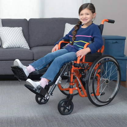 A young girl with a long brown braid, in a blue sweater and jeans, smiles while sitting in a Circle Specialty ZIGGO Pediatric Wheelchair in an orange color, with a gray couch and cushions visible in the living room background.