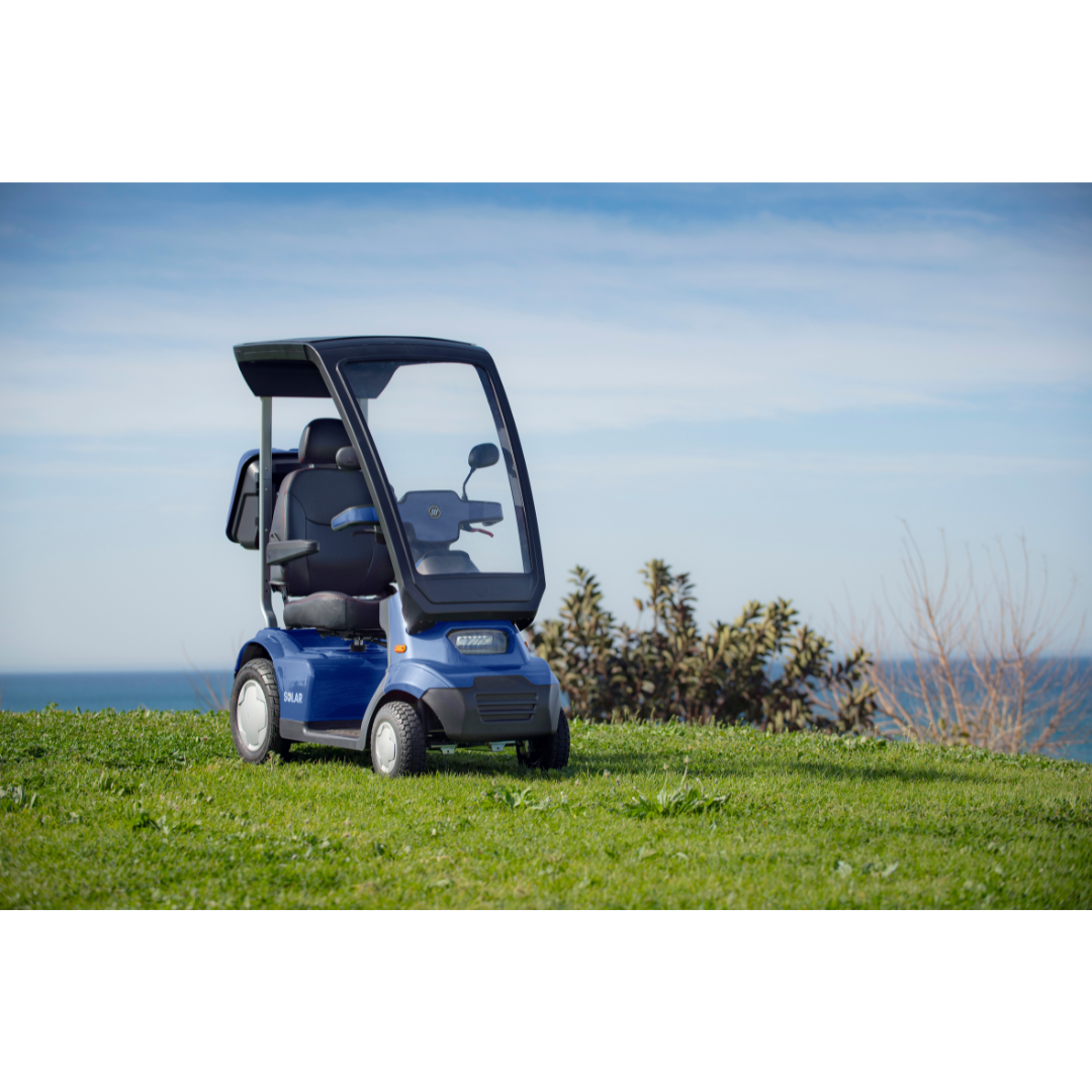 A small, blue Afikim Afiscooter Solar - Solar-Powered Bariatric Mobility Scooter with Canopy sits on a grassy hill by the ocean. The clear sky is dotted with a few clouds. Nearby bushes enhance its eco-friendly charm, portraying tranquility and cutting-edge mobility innovation.