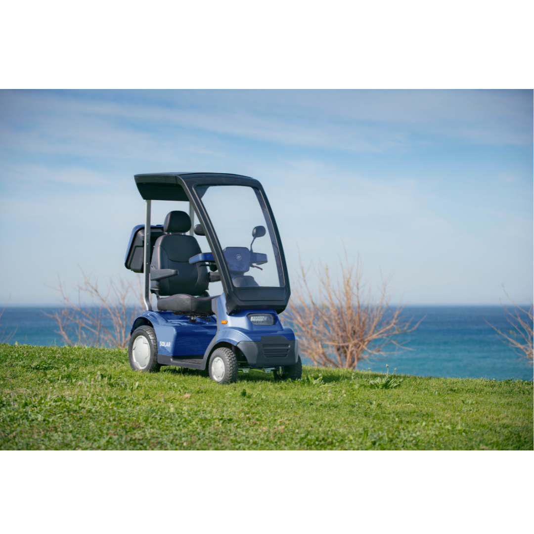 An Afikim Afiscooter Solar, a blue bariatric scooter with solar power and canopy, is parked on a grassy hill by the ocean. The partly cloudy sky and bare trees in the background add to the scenes tranquility. Its ergonomic tiller provides comfort while taking in the stunning views.