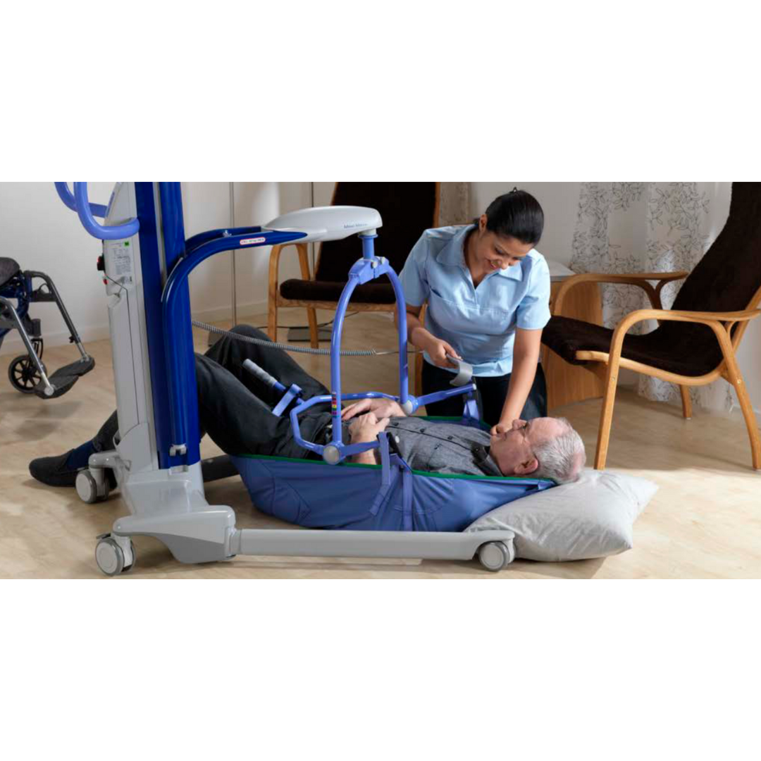 A caregiver demonstrates safe floor lift techniques using the ARJO Maxi Move Electric Floor Lifter & All-Purpose Patient Lift to assist an elderly man lying on a pillow, with a wheelchair and chairs visible in the background.