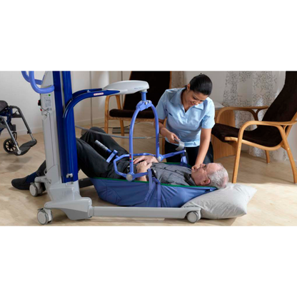 A caregiver demonstrates safe floor lift techniques using the ARJO Maxi Move Electric Floor Lifter & All-Purpose Patient Lift to assist an elderly man lying on a pillow, with a wheelchair and chairs visible in the background.
