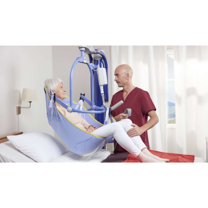 A healthcare worker assists an elderly woman seated in an ARJO 4-Point Spreader Sling for patient lifts beside a hospital bed in a bright room with white curtains.