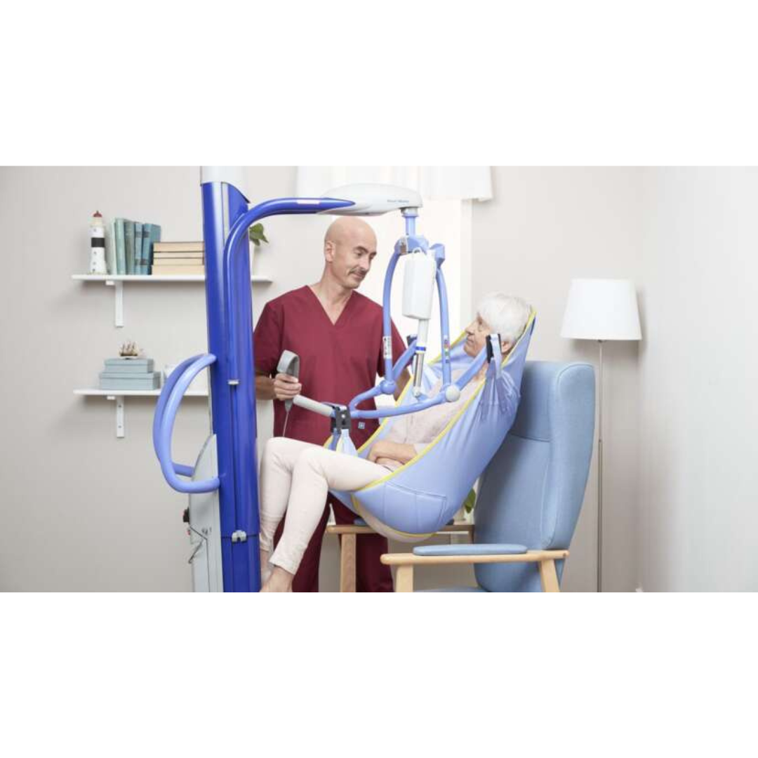 A healthcare worker in red scrubs uses the ARJO Maxi Move Electric Floor Lifter & All-Purpose Patient Lift to assist an elderly woman in a sling. They are in a bright, tidy room with bookshelves, a lamp, and a window.