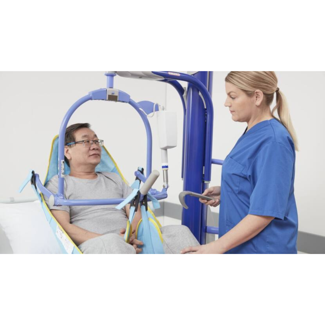 A healthcare worker in blue scrubs uses the ARJO Maxi Move Electric Floor Lifter & All-Purpose Patient Lift to assist a seated man in a gray shirt, secured in the sling, during transfer in a clinical setting.