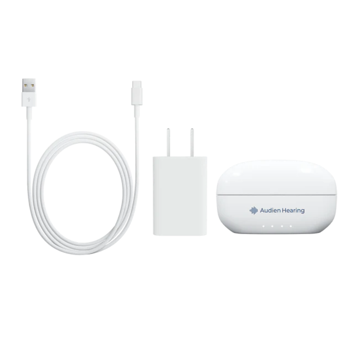 A white USB charging cable, a white wall charger, and a white Audien Hearing ION Pro Over-The-Ear High Def Hearing Aids case are arranged side by side on a plain background, designed for personalized hearing profiles.