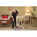 A man demonstrates the Bestcare SA Series Sit-To-Stand Compact Hydraulic Patient Lift for seamless patient transfers in a room with a hospital bed, chair, and lamp. The decor is warm with beige walls and a wooden floor, while a potted plant on a table adds greenery.