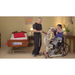 A man helps a woman in a wheelchair using the Bestcare BestStand™ SA400H/HE Mini Sit-To-Stand Patient Lift with 400lb capacity in a beige-walled room, showcasing a bed labeled Bestcare, an adjustable knee pad, and decor like a table with a lamp, painting, and plant.