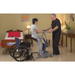 A caregiver helps a woman using the Bestcare STA450 Standing Transfer Aid in a bedroom with red-bedding bed, nightstand and lamp, nearby wheelchair for safety, plant, and decorative artwork.