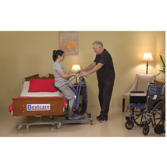 In a softly lit room, a caregiver helps a patient using the Bestcare STA450 Standing Transfer Aid, with essential equipment such as a bed, wheelchair, and lamps available. Patient safety is prioritized throughout the process.