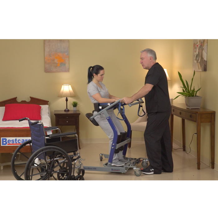 A caregiver skillfully operates the Bestcare STA450 Standing Transfer Aid near a bed beside a wheelchair. The room features beige walls, a painting, two lit lamps on wooden tables, and a brown bed with a red pillow displaying the Bestcare label.