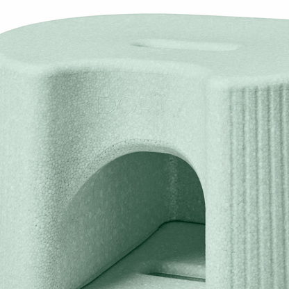 Close-up of a light green, textured byACRE DORICA foam block with a rounded edge, arch cutout, and top slot—serving as a versatile step stool or seat. Water-resistant, anti-slip surface is subtly embossed with the logo.