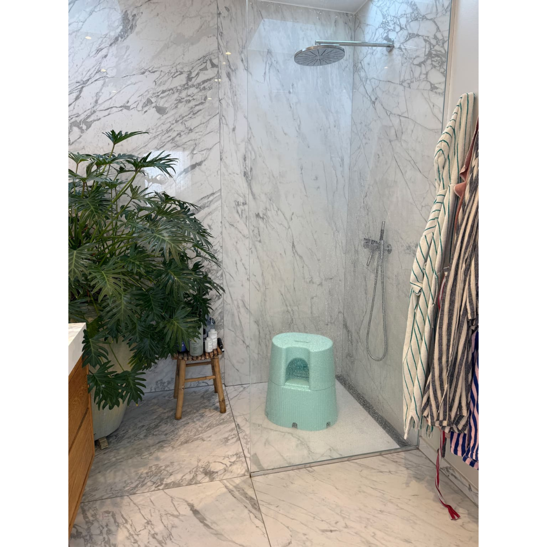 A modern marble-tiled bathroom with a walk-in shower, striped towels, a large potted plant, and a light blue byACRE DORICA Versatile Step Stool & Seat that’s water-resistant and anti-slip.