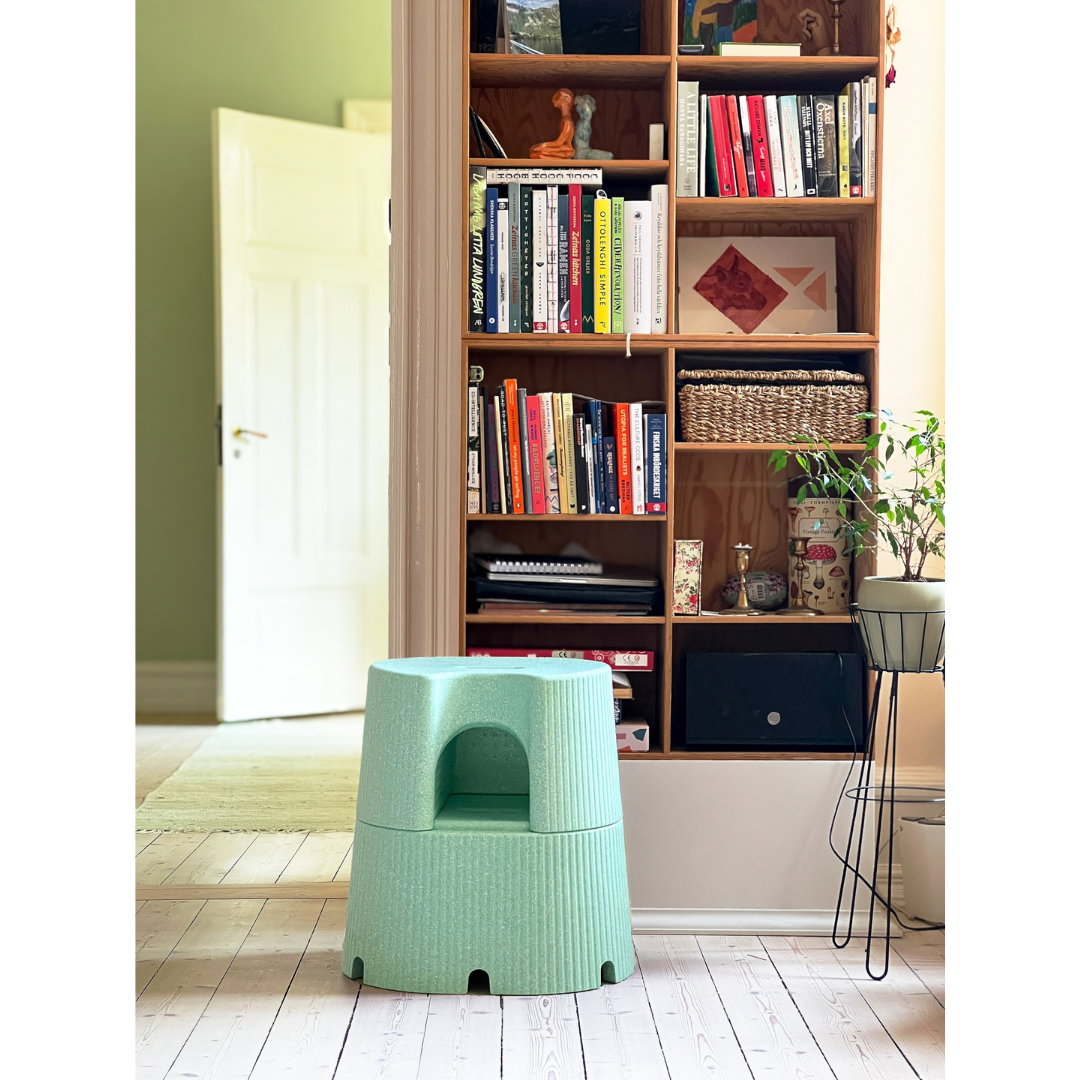 The byACRE DORICA Versatile Step Stool & Seat, in pale green with water-resistant and anti-slip features, sits on a wooden floor before a book-filled shelf. An open door and potted plant add charm to the bright, tidy room.