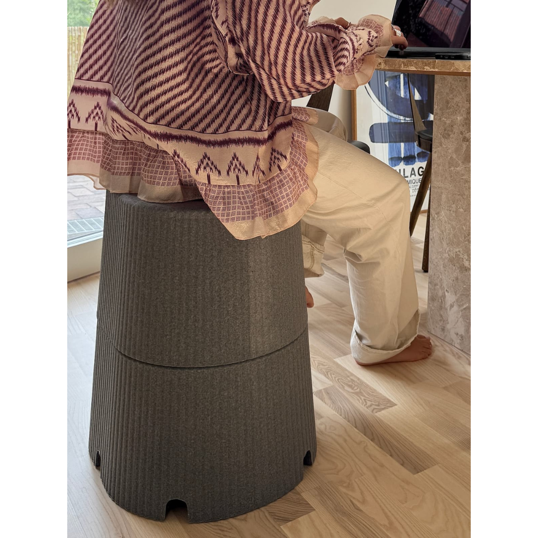 A person sits barefoot on the byACRE DORICA Versatile Step Stool & Seat, working at a laptop on a marble table in a bright room with wooden floors. The water-resistant, anti-slip stool adds comfort and functionality to the space.