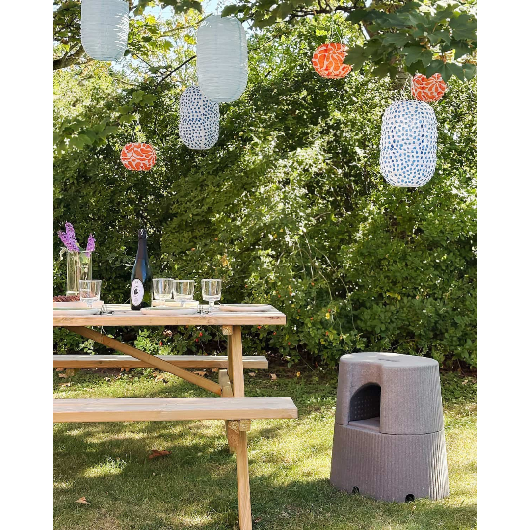 A wooden picnic table with glasses, plates, and a bottle sits on grass under paper lanterns, surrounded by greenery, with the byACRE DORICA Versatile Step Stool & Seat—water-resistant and anti-slip—placed nearby.