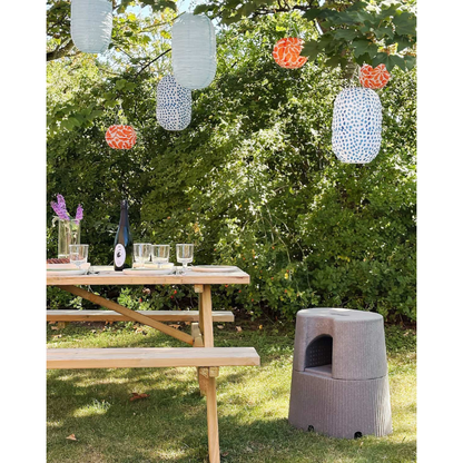 A wooden picnic table with glasses, plates, and a bottle sits on grass under paper lanterns, surrounded by greenery, with the byACRE DORICA Versatile Step Stool & Seat—water-resistant and anti-slip—placed nearby.