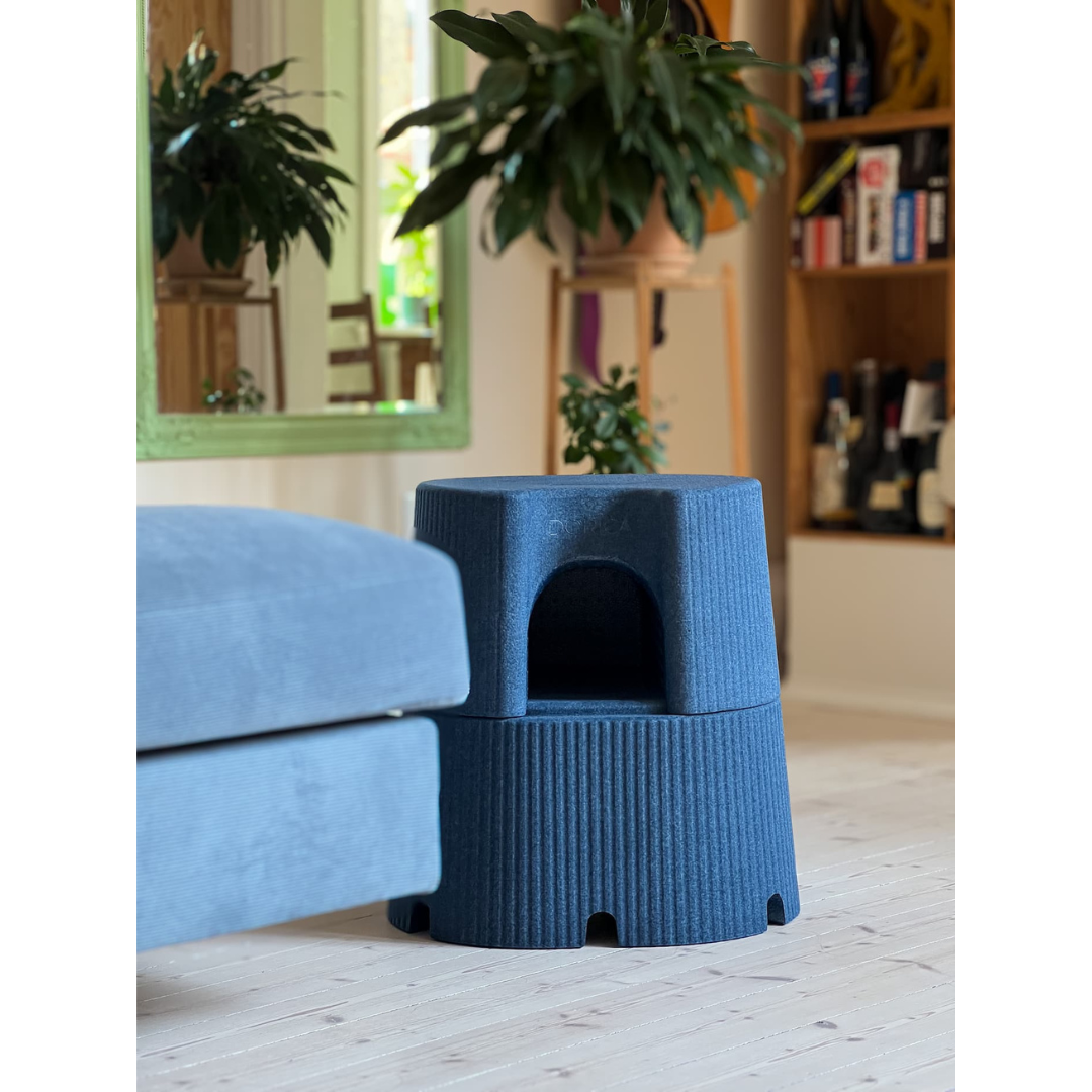 A blue textured cat house and a matching blue sofa sit beside the byACRE DORICA Versatile Step Stool & Seat—water-resistant and anti-slip—in a cozy living room, with plants, a mirror, and shelves of bottles and board games in the background.