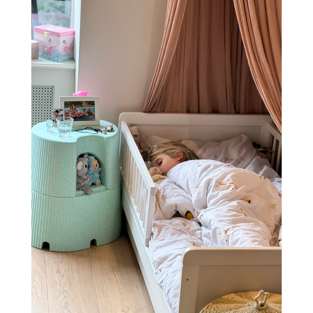 A young child sleeps in a white bed with animal-patterned bedding and a beige canopy. Next to the bed, the byACRE DORICA Versatile Step Stool & Seat holds a glass of water, framed photos, and stuffed animals in its lower cubby.