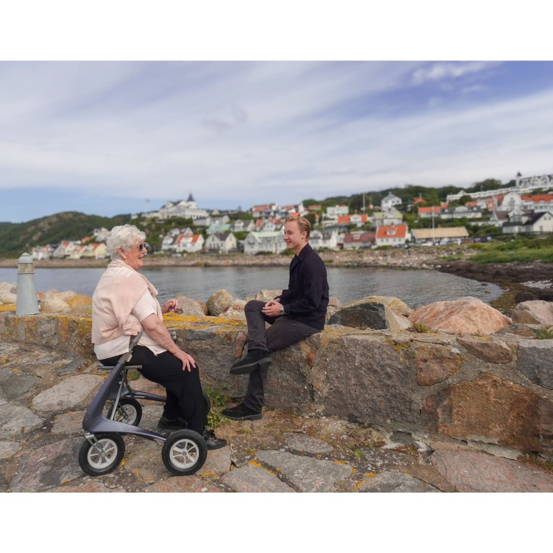 An elderly woman with a ByACRE Carbon Overland All-Terrain Rollator and her companion sit on a stone wall by the water, chatting as colorful houses and green hills stretch behind them beneath a partly cloudy sky.