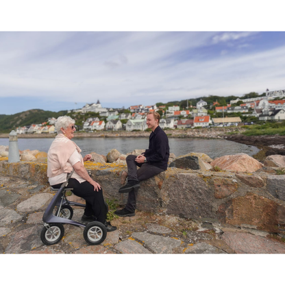 An elderly woman with a ByACRE Carbon Overland All-Terrain Rollator and her companion sit on a stone wall by the water, chatting as colorful houses and green hills stretch behind them beneath a partly cloudy sky.