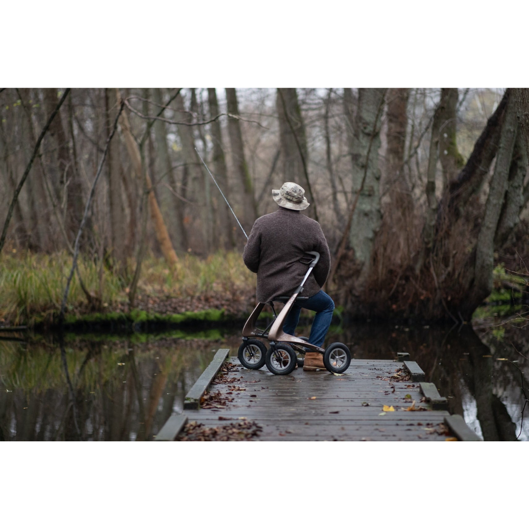 An older person in a hat and coat sits on their ByACRE Carbon Overland All-Terrain Rollator, fishing from a wooden dock in a peaceful, leaf-strewn forest beside calm water.