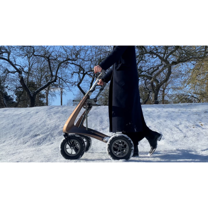 Clad in a long dark coat, a person navigates snowy terrain beneath leafless trees on a sunny day, relying on the ByACRE Carbon Overland All-Terrain Rollator as their sturdy four-wheeled companion.