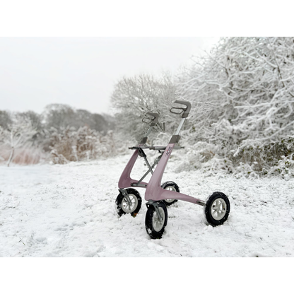 The ByACRE Carbon Overland All-Terrain Rollator, with its pink frame and four wheels, stands on a snowy path, ready for winter adventures among snow-covered trees and bushes.