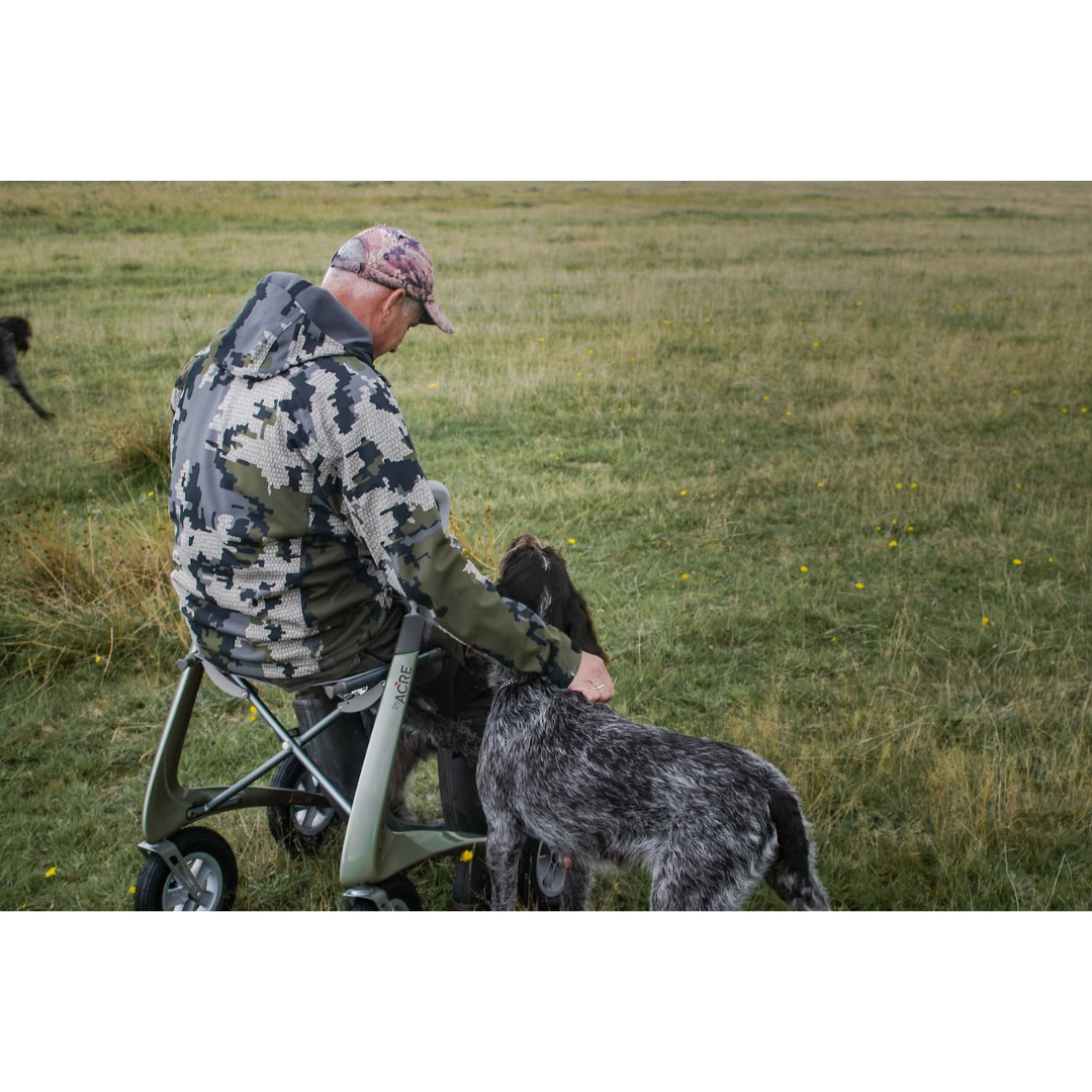 An older man in a camouflage jacket and cap sits on his ByACRE Carbon Overland All-Terrain Rollator in a grassy field, petting his two black and white dogs standing before him—his loyal adventure companions.