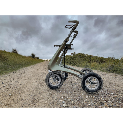 The ByACRE Carbon Overland All-Terrain Rollator—a modern four-wheeled companion—is parked on a gravel path outdoors, surrounded by green grass and bushes beneath a cloudy, overcast sky.