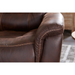 Close-up of the brown leather Golden Tech PR446 DeLuna™ Dione Recliner, showcasing visible stitching and highlighting the armrests craftsmanship. A wooden table and rug blur in the background. Enjoy ultimate relaxation with MaxiComfort Positioning settings.