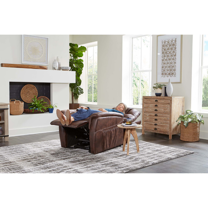 A person relaxes in a cozy living room on a Golden Tech PR446 DeLuna™ Dione Recliner with Lift Assist - Medium, amidst wooden furniture, potted plants, and a side table. A fireplace adds warmth while large windows provide natural light over the geometric patterned rug.