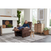 A woman relaxes in a brown Golden Tech PR446 DeLuna™ Dione Recliner with Lift Assist, enhancing her cozy living room. The space includes a patterned rug, wooden side table with a plant, chest of drawers, decorative wall art, and fireplace, with large windows filling the room with natural light.