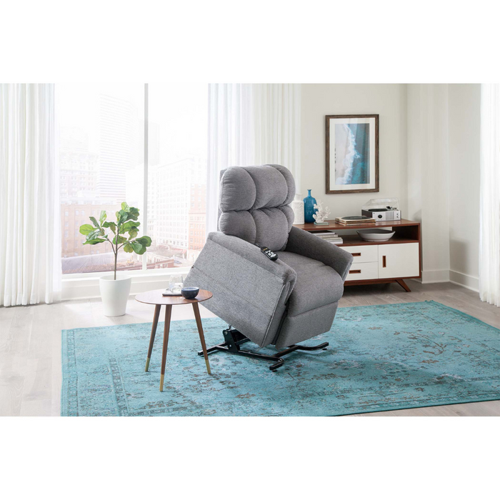 In a modern living room, the Golden PR535 MaxiComfort Recliner in gray is positioned upright for ergonomic support next to a small round table and potted plant. A blue patterned rug adorns the floor, with a sideboard and decor against the wall, while large windows showcase an urban cityscape.