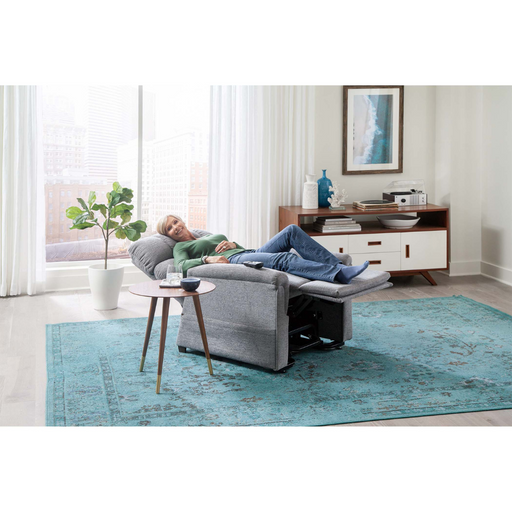 A person unwinds on a Golden PR535 MaxiComfort Recliner in their modern living room, wearing a green sweater and jeans. The room has large windows, a blue rug, a side table with a book, and decorative items on the console from the Comforter Series.