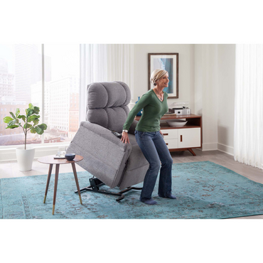 A woman uses the Golden PR531 Comforter Recliner w/ Lift Assist in a bright living room with a blue rug, round side table, plant, and a cabinet with electronics and framed artwork, enjoying customized comfort.