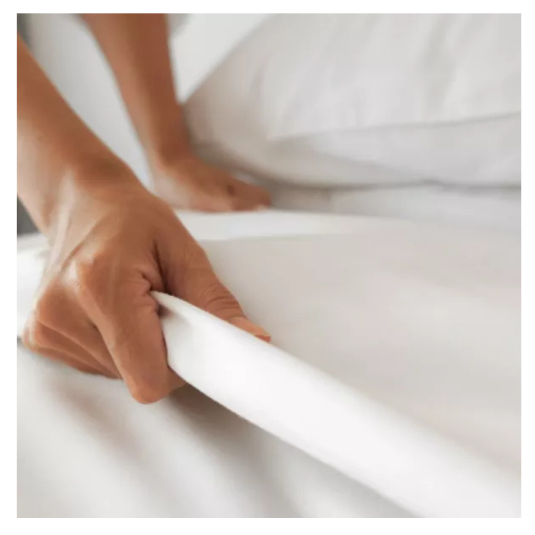 A close-up of a hand tucking iCare Medical Mattress Bed Sheets, made from 100% cotton, onto an adjustable bed mattress with a white pillow in the background, showing a neatly made bed with premium linen.