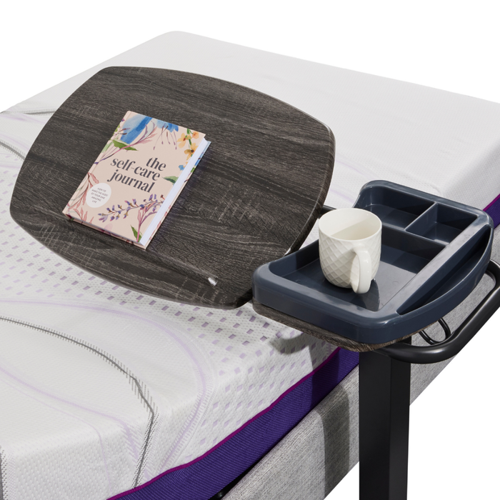 An iCare Medical Over Bed Tilting Table with a gray adjustable tray holds The Self-Care Journal and a blue cup holder with a white textured mug. The mattress, with white and purple accents, creates a cozy, functional space.