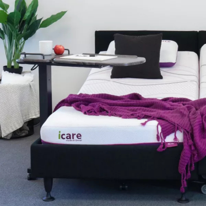 The iCare Medical Over Bed Tilting Table - Height Adjustable, shown beside an adjustable iCare bed with white and purple bedding, holds a mug and apple, while a plant and towels in the background highlight comfort and mobility.