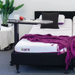 The iCare Medical Over Bed Tilting Table - Height Adjustable, shown beside an adjustable iCare bed with white and purple bedding, holds a mug and apple, while a plant and towels in the background highlight comfort and mobility.