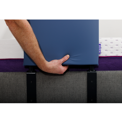 A person’s hand holds the iCare Medical Bed Safety Rail, a sturdy blue support attached to a bed with a gray upholstered base and white mattress for fall prevention.