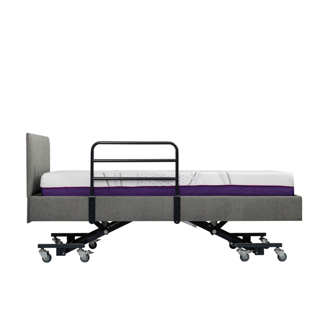 Side view of an adjustable hospital-style bed with wheels, gray upholstered frame, vertical headboard, and an iCare Medical Bed Safety Rail for fall prevention. The mattress features a white top and a purple layer underneath.